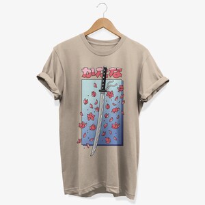 May include: A beige t-shirt featuring a graphic of a katana sword with pink cherry blossoms falling around it. The Japanese text "かわええ" is printed above the sword.