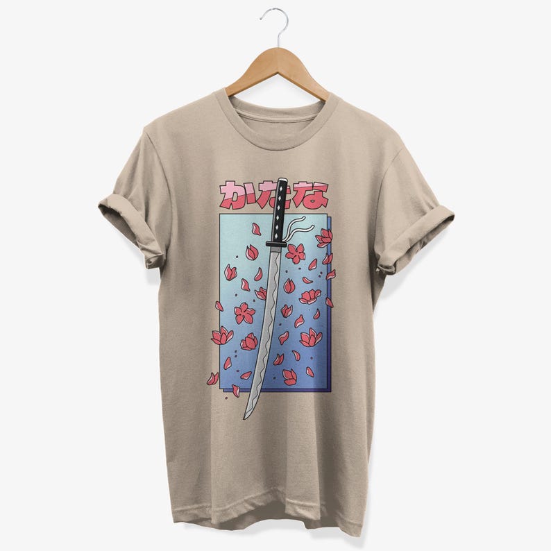 May include: A beige t-shirt featuring a graphic of a katana sword with pink cherry blossoms falling around it. The Japanese text "かわええ" is printed above the sword.
