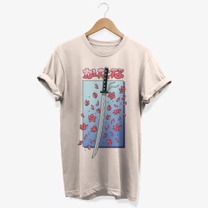 May include: A beige t-shirt featuring a graphic of a katana sword with pink cherry blossoms falling around it. The Japanese text on the shirt reads "カワイイ"