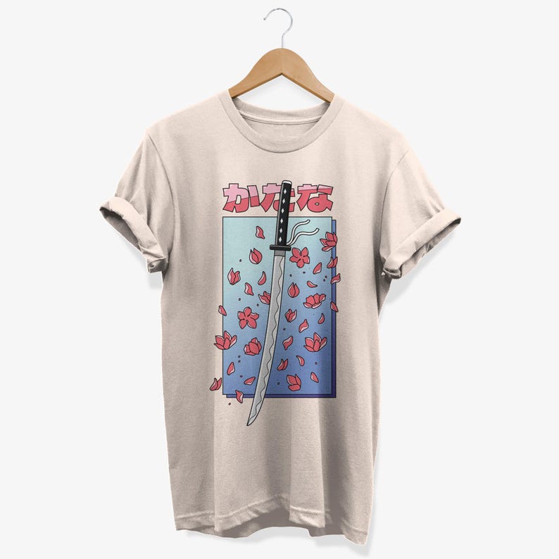 May include: A beige t-shirt featuring a graphic of a katana sword with pink cherry blossoms falling around it. The Japanese text on the shirt reads "カワイイ"