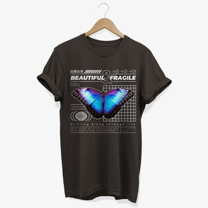 May include: A dark gray t-shirt with a graphic design featuring a blue and purple butterfly on a grid background. The text "BEAUTIFUL & FRAGILE" is above the butterfly, and the text "drifting along through life" is below the butterfly.