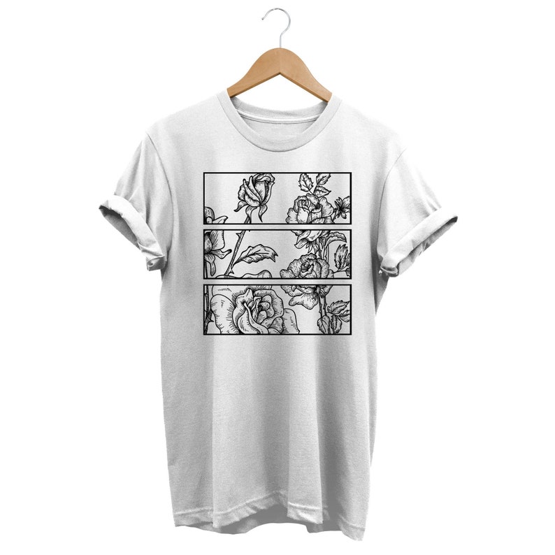 May include: White t-shirt featuring a black and white floral design. The design is divided into three horizontal rectangular panels, each showcasing detailed rose illustrations. The shirt is displayed on a wooden hanger.