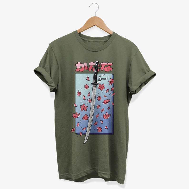 May include: A green t-shirt featuring a graphic of a katana sword with pink cherry blossoms falling around it. The Japanese text on the shirt reads "カワイイ"