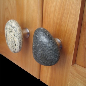 May include: Two cabinet knobs made from natural stones. One knob is a light gray and white stone with a smooth, rounded surface. The other knob is a dark gray stone with a rough, textured surface.