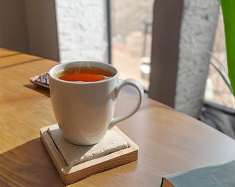 May include: A white ceramic mug filled with amber-coloured tea, resting on a square stone coaster with a light-coloured wooden frame. The mug features a curved handle and a smooth finish, while the coaster displays a porous, textured surface.