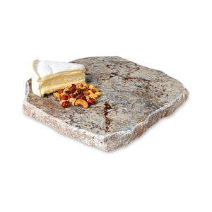 May include: A square, light brown granite serving board with a rough, uneven edge. A wedge of white brie cheese and a scattering of mixed nuts are on the board.