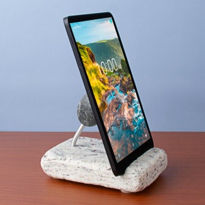 May include: A black tablet with a landscape image of a river and mountains on the screen. The tablet is propped up on a white stone stand with a round stone on the front.