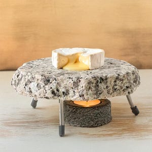May include: A round granite cheese warmer with a metal stand and a small candle holder. A round wheel of brie cheese is melting on top of the warmer.