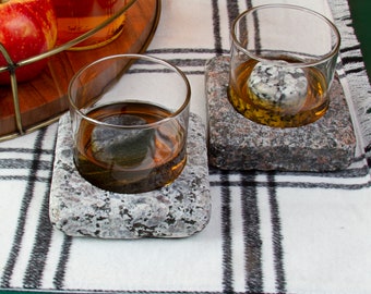 May include: Two clear glass tumblers, each containing a round granite stone, are set into square granite coasters with a speckled gray and black texture. The glasses are filled with an amber-colored liquid and rest on a white plaid cloth.