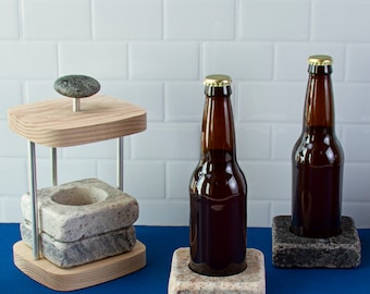 May include: Two brown glass beer bottles with gold caps are sitting on granite coasters. The coasters are on a blue surface. One bottle is on a wooden stand with a granite coaster.