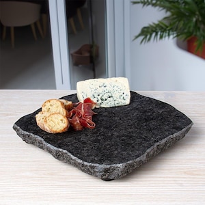 May include: A square, dark gray granite serving board with a rough, natural chiseled edge. The stone platter holds a wedge of blue-veined cheese, several thin slices of cured meat, and three round pieces of toasted bread arranged on the surface.