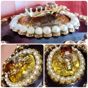 K&ouml;nnte beinhalten: Vintage goldfarbene Brosche mit einem gro&szlig;en bernsteinfarbenen Stein, umgeben von kleinen Perlen. Ein detailliertes Insektenmuster mit einem roten Edelstein sitzt auf dem bernsteinfarbenen Stein. Die Brosche hat einen dunklen Hintergrund.
