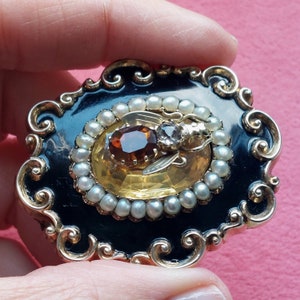 K&ouml;nnte beinhalten: Eine verzierte Vintage-Brosche mit schwarzem Emaille-Sockel und goldfarbenem Filigran. Die Brosche hat einen ovalen, bernsteinfarbenen Stein, umgeben von kleinen Perlen und einem dekorativen, insektenf&ouml;rmigen Element. Das Design ist elegant und detailliert.