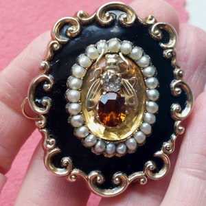 K&ouml;nnte beinhalten: Vintage Brosche in Goldoptik mit schwarzem Emaille-Hintergrund und ovaler Mitte. Die Mitte zeigt ein Bienenmuster mit einem gro&szlig;en bernsteinfarbenen Stein, einem klaren Stein und kleinen Perlen. Die Brosche hat einen verzierten, gerollten Rand.