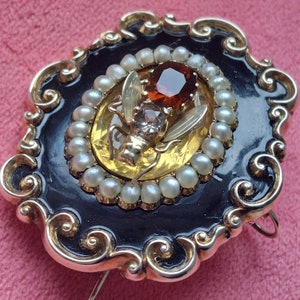 K&ouml;nnte beinhalten: Verzierte Brosche mit goldfarbenem Rahmen und schwarzem Emaille-Hintergrund. Die Brosche hat ein ovales Mitteldesign mit einem gro&szlig;en, facettierten, rotbraunen Edelstein, umgeben von kleinen Perlen. In der Mitte befindet sich eine goldfarbene Fliege mit Edelsteinakzenten.