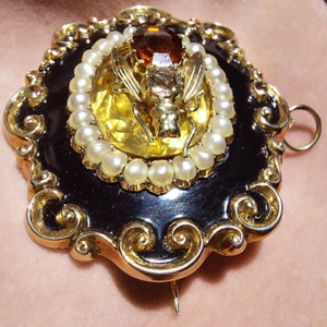 K&ouml;nnte beinhalten: Vintage Brosche in Goldoptik mit schwarzem Emaille-Rahmen und aufwendigen Golddetails. Die Brosche zeigt ein zentrales Design einer goldenen Biene, umgeben von Kunstperlen und einem gro&szlig;en bernsteinfarbenen Stein. Ein kleinerer klarer Stein befindet sich &uuml;ber der Biene.