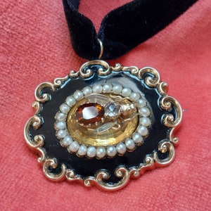 K&ouml;nnte beinhalten: Eine Halskette mit Anh&auml;nger im Vintage-Stil mit einem ovalen Anh&auml;nger aus schwarzem Emaille und einem goldfarbenen Filigranrahmen. Der Anh&auml;nger ist mit einem bernsteinfarbenen Mittelstein, kleinen Perlen und einem goldfarbenen Insektenmuster verziert, das an einem schwarzen Samtband h&auml;ngt.