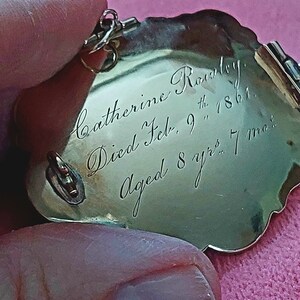 K&ouml;nnte beinhalten: Ein goldfarbenes, herzf&ouml;rmiges Medaillon mit graviertem Text. Der Text lautet "Catherine Rowley. Gestorben am 9. Februar 1861. Alter 8 Jahre 7 Monate." Das Medaillon hat einen dekorativen Rand und einen kleinen Verschluss.
