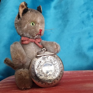May include: A plush gray cat toy holding a silver locket with an ornate design. The locket has a hinged lid and a detailed floral pattern.