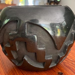 May include: A black, round ceramic bowl with a glossy finish. The bowl features a repeating geometric pattern carved into its surface, creating a textured design. The bowl sits on a wooden surface.