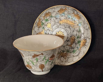 May include: A white porcelain teacup and saucer set with a gold rim. The cup and saucer are decorated with a hand-painted design of fish, crabs, and bamboo in gold and green.
