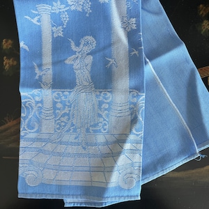 Puede incluir: Una toalla de té de lino azul con un diseño bordado blanco de una mujer de pie en un jardín. El diseño incluye columnas, pájaros y follaje.