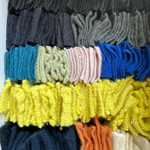 Op de afbeelding: Een verzameling kleurrijke, getextureerde stofstalen gerangschikt in rijen. De stalen tonen een verscheidenheid aan kleuren, waaronder zwart, marineblauw, grijs, blauwgroen, geel, oranje en crème. De stof lijkt een geluste of getextureerde brei te zijn.