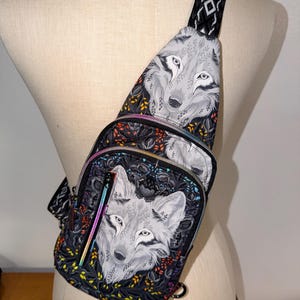 May include: A black sling bag featuring a detailed wolf illustration. The bag has a rainbow zipper and a patterned strap. The wolf design is set against a backdrop of dark floral patterns with accents of orange, yellow, and green.