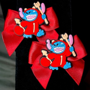 May include: Two red hair bows with blue and white cartoon Stitch characters wearing red devil costumes. The Stitch characters are holding pitchforks.