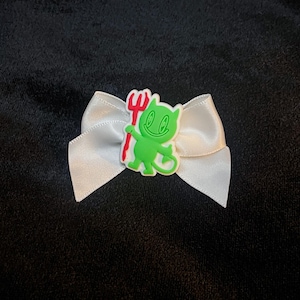 Puede incluir: Un lazo de satén blanco con un personaje de diablo verde sosteniendo un tridente rojo. El diablo tiene una cara sonriente y una cola larga. El lazo está sobre un fondo negro.