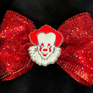 Könnte beinhalten: Eine rote Glitzer-Schleife mit einem weißen und roten Clown-Gesicht in der Mitte. Die Schleife besteht aus einem glänzenden, strukturierten Material und ist wahrscheinlich ein Haaraccessoire oder ein dekorativer Artikel. Das Clown-Gesicht hat ein weißes Gesicht, rotes Haar und eine rote Nase.