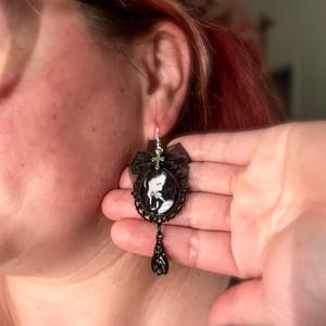 May include: Black and white cameo earrings with a black bow and a dangling teardrop charm. The cameo features a woman with a cross.
