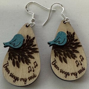 May include: Wooden teardrop earrings with a light wood grain finish. Each earring features a teal bird perched on a branch design, with the words "Then sings my soul" and musical notes engraved on the wood. Silver-tone ear wires.