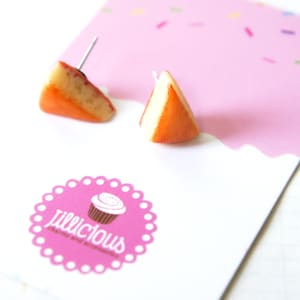 May include: A pair of stud earrings shaped like small orange and white triangular cheese wedges. The earrings are attached to a white display card featuring a pink top section with sprinkles and a circular logo that reads Jillicious.