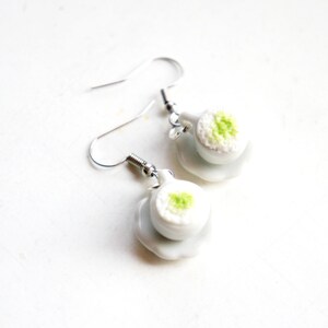 May include: A pair of white ceramic earrings shaped like a small bowl of rice with a green garnish. The earrings are on a silver hook.