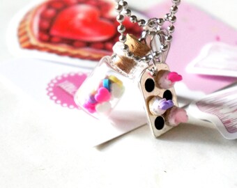 May include: A silver chain necklace with two charms: a small glass bottle filled with colorful sprinkles and a silver cupcake baking pan with three miniature cupcakes. The cupcakes are decorated with pink, purple, and white frosting.