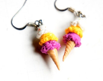 May include: A pair of dangle earrings featuring a three-scoop ice cream cone with white, yellow, and purple scoops. The cone is made of polymer clay and is attached to silver-toned earring hooks.