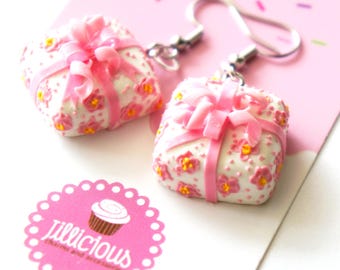 May include: A pair of white earrings shaped like gift boxes, adorned with pink bows and floral accents. The earrings have silver-colored hooks and are set against a pink background. The logo "jillicious charms and accessories" is visible.