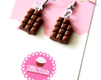 May include: A pair of chocolate bar earrings with silver-colored hooks. The earrings are a rich brown color and are displayed on a pink card with the text "Jillicious charms and accessories" and a cupcake logo.