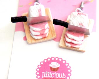 May include: A pair of novelty earrings featuring miniature wooden cutting boards with white and pink food, a silver knife, and a white and pink food item. The earrings have silver hooks. The earrings are on a pink background with a pink logo that says "Jillicious charms and accessories."