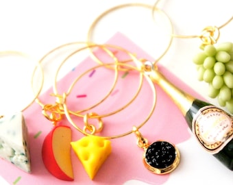 May include: Set of four gold wine glass charms with different food charms. The charms include a wedge of cheese, a slice of apple, a small dish of caviar, and a miniature bottle of champagne.