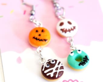 May include: A pair of Halloween-themed dangle earrings. One earring features a pumpkin, a skull, and a chocolate donut with white icing. The other earring has a pumpkin, a skull, and a teal donut with black icing. The earrings are connected with silver-colored chains.