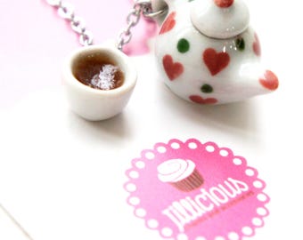 May include: A miniature teapot and teacup necklace. The white teapot is decorated with red hearts and green dots. The teacup is filled with a brown liquid. The necklace chain is silver. A pink sticker with the word "jillicious" is in the image.