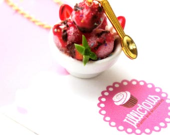 May include: A miniature dessert charm necklace featuring a small white bowl filled with pink ice cream scoops, drizzled with chocolate sauce and topped with red strawberry slices. A gold-colored spoon rests beside the bowl. The charm is attached to a gold chain.
