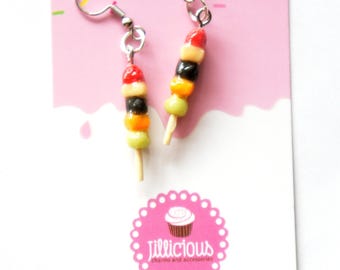 May include: A pair of dangle earrings shaped like miniature fruit skewers. Each earring features a wooden stick with colorful beads resembling fruit slices in red, white, black, orange, and green. The earrings hang from silver-colored fish hook ear wires.