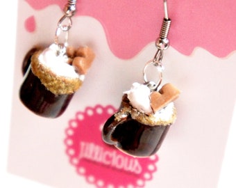 May include: A pair of dangle earrings featuring miniature mugs filled with hot chocolate, topped with whipped cream and chocolate sprinkles. The mugs are brown with gold glitter around the rim. The earrings are silver.