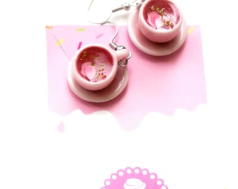 May include: Pink teacup earrings with gold glitter and flower accents. Each miniature cup sits on a matching saucer, suspended from silver-tone ear wires. The earrings are displayed on a pink background with the word "Tillicious" at the bottom.