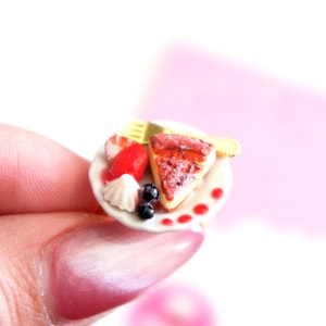 Peut inclure: Petite assiette de dessert avec une part de cheesecake, une fraise, de la crème fouettée et des myrtilles. Une fourchette et un couteau jaunes sont sur l'assiette. L'assiette a des points rouges sur le bord. Le dessert est sur une assiette blanche.