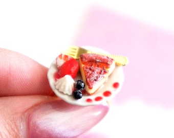 May include: Miniature dessert plate with a slice of cheesecake, a strawberry, whipped cream, and blueberries. A yellow fork and knife are on the plate. The plate has red dots around the edge. The dessert is on a white plate.