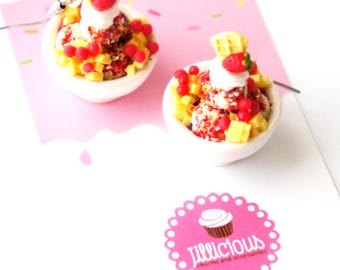 May include: Two miniature sundae earrings. Each sundae is in a white bowl and topped with whipped cream, a strawberry, and a waffle piece. The sundae has red and yellow fruit pieces and sprinkles. The background is pink with a logo that says "Jillicious charms and accessories."